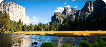 A panoramic view of Yosemite Valley with the Merced River flowing in the foreground, bordered by a field of golden grass. The massive granite face of El Capitan is on the left, and other rugged mountain peaks are on the right, with a waterfall visible in the distance.