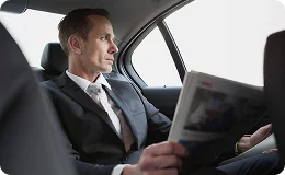 A businessman in a suit reads a newspaper in the back seat of a car, looking out the window.