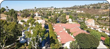 Aerial view of a residential neighborhood on a hillside, featuring buildings with terracotta roofs surrounded by lush green trees and shrubs. The terrain slopes upward in the background.