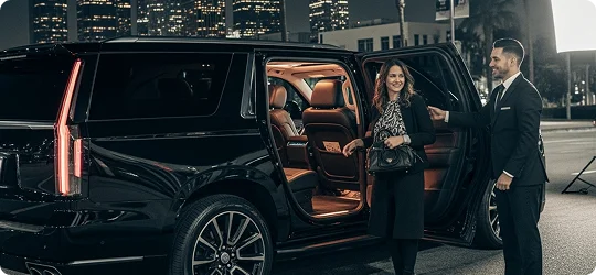 A male chauffeur in a suit opens the door of a black Cadillac Escalade SUV for a female passenger at night.