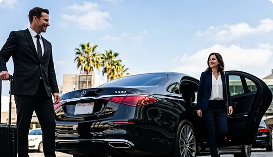 A smiling chauffeur in a dark suit stands beside rolling luggage next to a black luxury sedan. A businesswoman steps out of the open rear passenger door under a sunny sky with palm trees in the background.