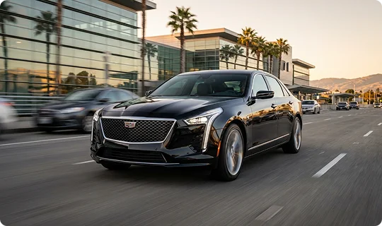 A black Cadillac sedan driving on a multi-lane city road during golden hour. The car is in sharp focus in the foreground, while the background features modern glass buildings and palm trees with a slight motion blur.