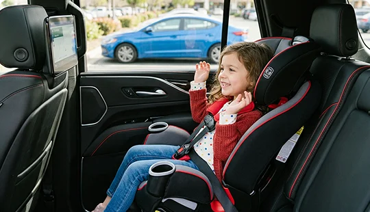 A young smiling girl is buckled into a secure, black and red child safety car seat installed in the back of a luxury vehicle. The vehicle features premium black leather seats with red stitching and an entertainment screen mounted on the back of the front headrest.