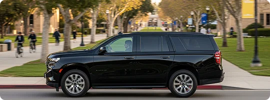 A black Chevrolet Tahoe SUV is driving along a tree-lined avenue on a sunny day. The scene appears to be on a large university campus, with students riding bicycles and walking on the sidewalks in the blurred background.