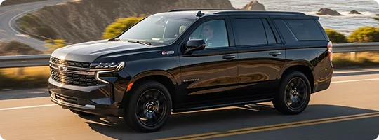A sleek, black Chevrolet Tahoe SUV is driving on a two-lane road that winds along a scenic, hilly coastline. The SUV is shown from a low front three-quarter angle in bright sunlight.