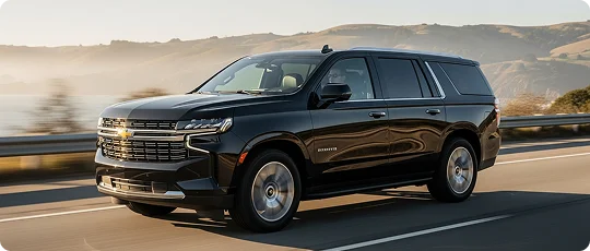 A shiny black Chevrolet Tahoe SUV is driving along a scenic coastal highway under a bright, slightly hazy sky. Rolling green-brown hills and a body of water are visible in the background.