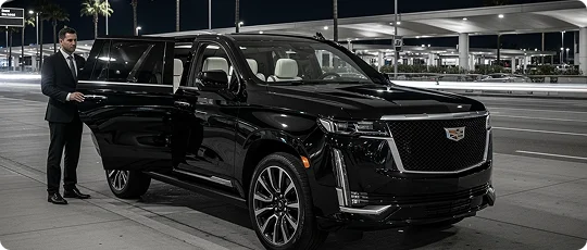 A male chauffeur in a suit stands next to a black Cadillac Escalade SUV with the door open. The scene is an airport pickup at night, with streetlights and palm trees in the background.