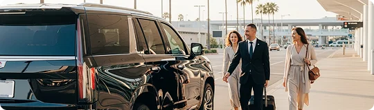 A chauffeur in a suit assisting two passengers with their luggage near a black SUV at an airport.