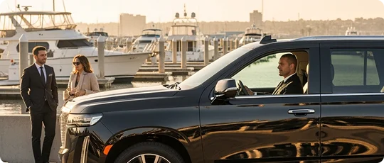 A chauffeur drives a black Cadillac Escalade SUV past a yacht harbor where a formally dressed couple is standing.