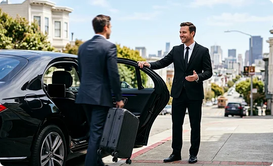 A smiling chauffeur in a dark suit opens the door of a black luxury car on a city street. A male passenger pulling a black suitcase walks toward the open car door with a cityscape in the background.