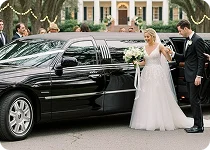 A newly married couple, the bride in a white gown and the groom in a tuxedo, stand next to a black stretch limousine outside a formal building.