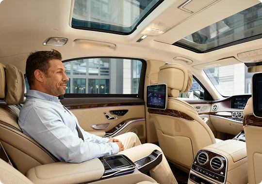 Smiling man seated in a spacious tan leather car interior with dual sunroofs, backseat entertainment screens, and extended footrests, highlighting premium comfort.