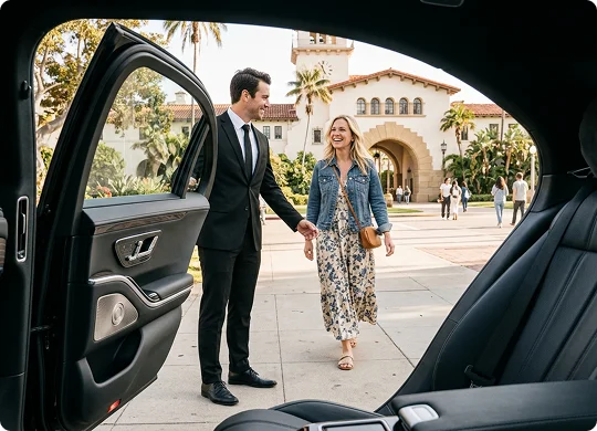 A smiling chauffeur in a suit holds open the rear door of a luxury car for a blonde woman in a denim jacket and patterned dress. The perspective from inside the car frames the pair on a sunny plaza in front of a historic Spanish-style building with a clock tower.