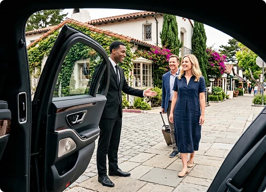 A smiling chauffeur in a suit holds open the door of a luxury car while gesturing to welcome a couple. The perspective from inside the vehicle shows a man rolling a suitcase behind a blonde woman in a navy dress on a cobblestone street.