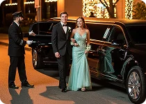 A chauffeur in a suit and cap holds the door of a black luxury vehicle for a smiling couple dressed formally, the woman wearing a mint-green gown.