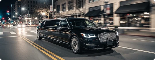 A black stretch limousine is driving quickly down a city street at night. The motion blur captures the speed of the vehicle as it drives past storefronts and streetlights.
