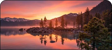 A mesmerizing view of Lake Tahoe with orange sunset behind the lake mountains, rocks, pine trees and calm lake water visible.
