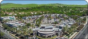 An aerial view of a large outdoor shopping center with extensive parking, palm trees, and surrounded by residential areas and green, rolling hills.
