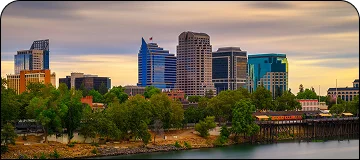 Scenic cloudy view of Downtown Sacramento with visible skyscrapers and bushy green trees with lake visible below.
