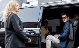A woman stands outside a black luxury van as a man wearing sunglasses sits inside the vehicle, looking at the camera.