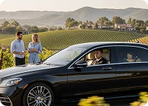 A black Mercedes Benz S class with a chauffeur seated inside is parked in a lush green rural area with fields, luxury condos, and green mountains visible in the background. A happy young couple stands right besides the car.