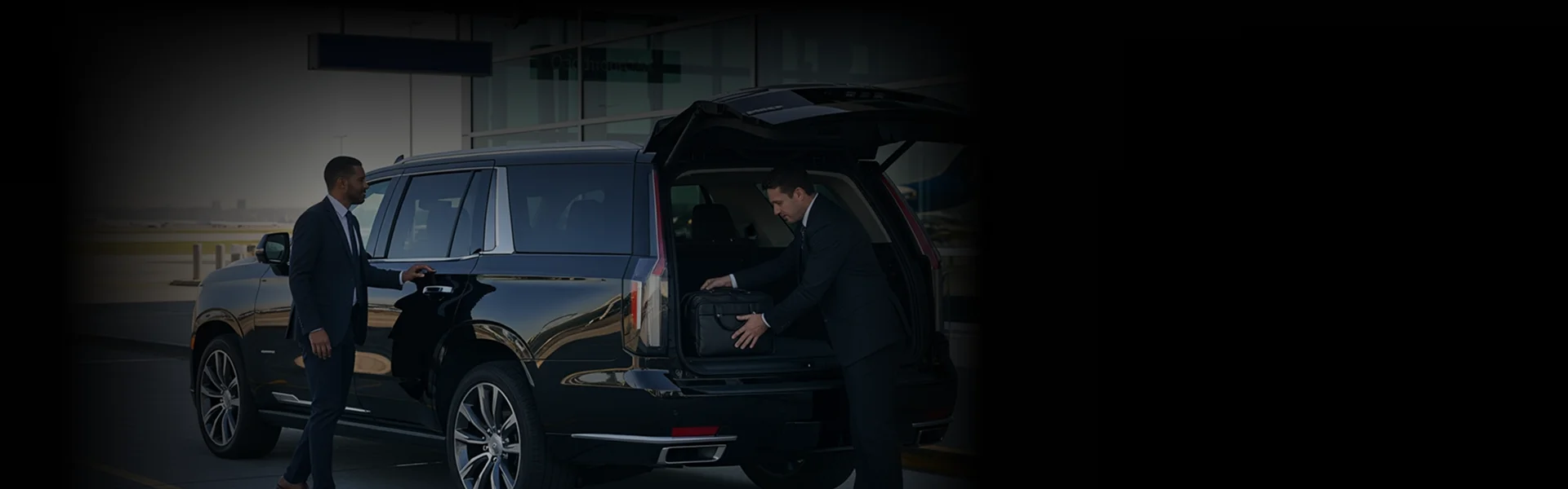 Two men in suits are standing next to a black Cadillac Escalade SUV outside at an airport or terminal. The passenger is standing by the rear passenger door, while the chauffeur places a black briefcase into the vehicle's open trunk. The scene is dark, with an urban skyline visible in the distance.