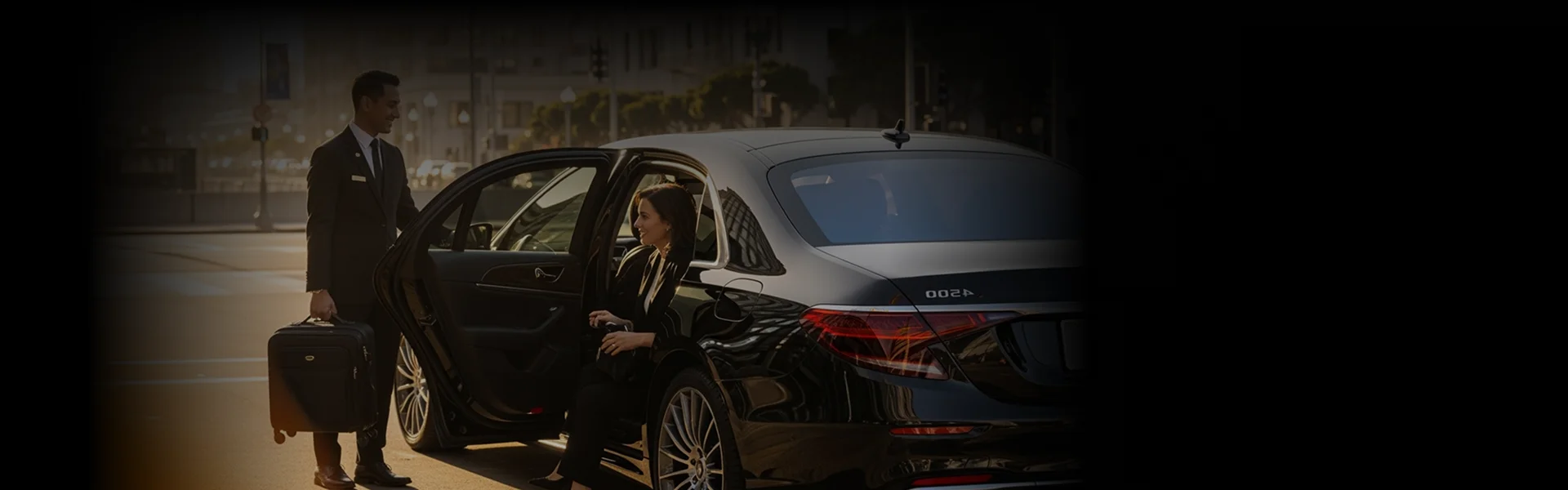 A professionally dressed chauffeur holds open the rear door of a black luxury sedan for a smiling woman. The chauffeur is also holding a rolling suitcase on a city street.