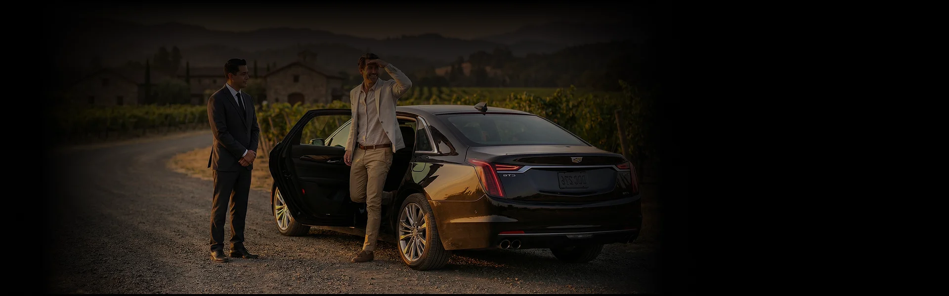 A chauffeur in a suit stands by the open door of a black sedan as a man in a tan jacket and pants steps out and shields his eyes from the sun.