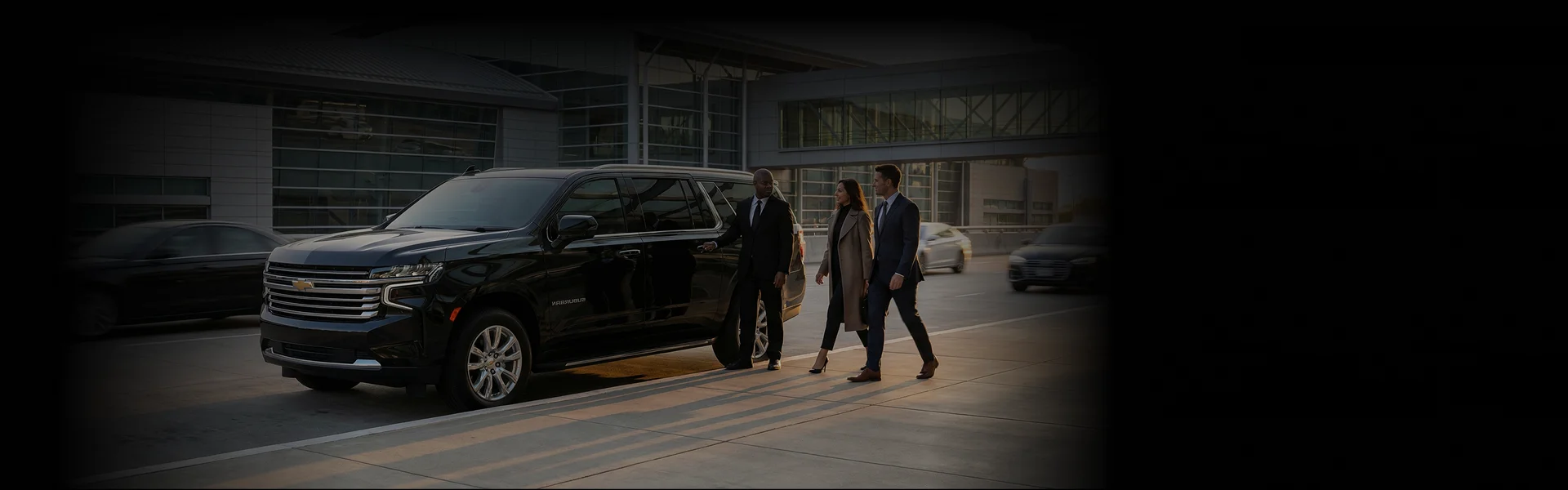 A decently dressed couple walks towards a black Chevrolet Suburban parked on the side lane with a chauffeur standing besides it. Background lightly shows a glass building and a car.