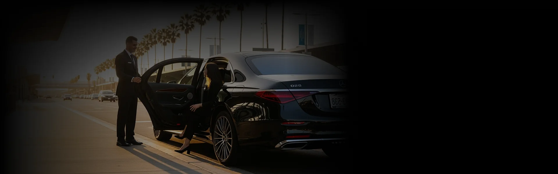 A chauffeur in a suit holds the door open for a woman getting into a black luxury sedan on a palm-lined city street.