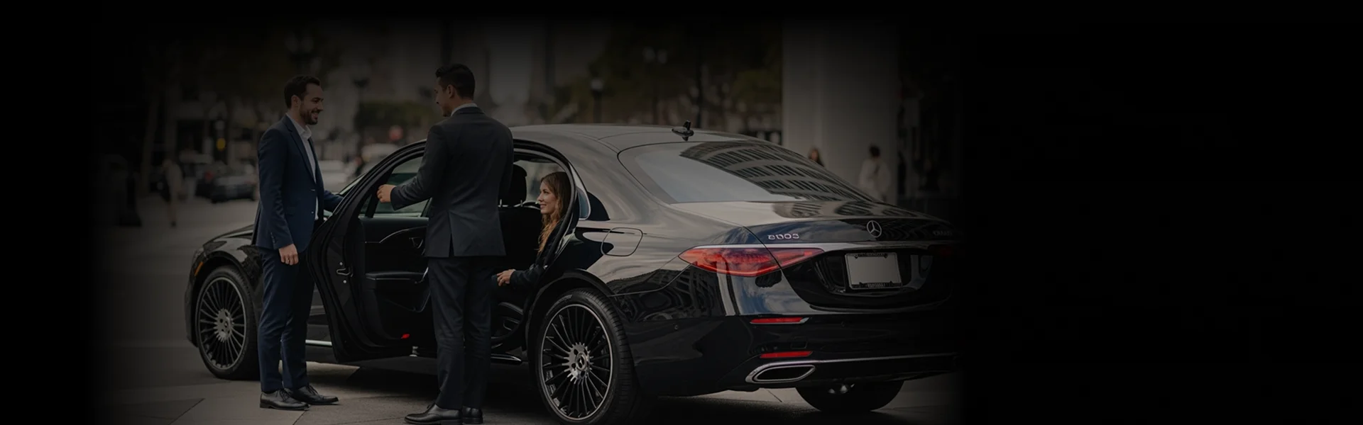 A chauffeur in a suit holds the door open for a woman seated in a luxurious black sedan, while a male passenger stands outside smiling.