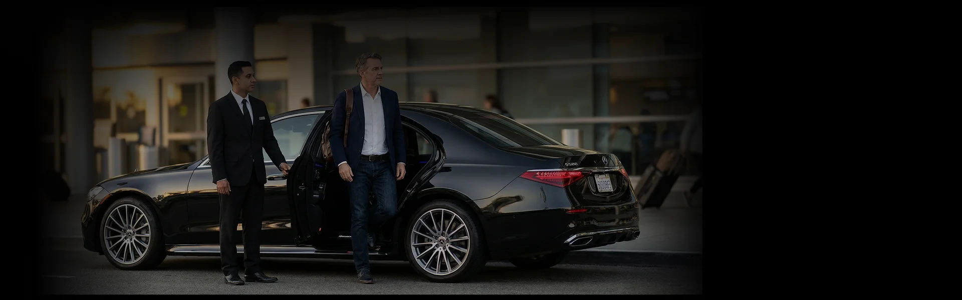 A middle-aged male passenger exiting a black business sedan (probably a Mercedes-Benz S-Class) while a black suited chauffeur hold the rear door opened for him.